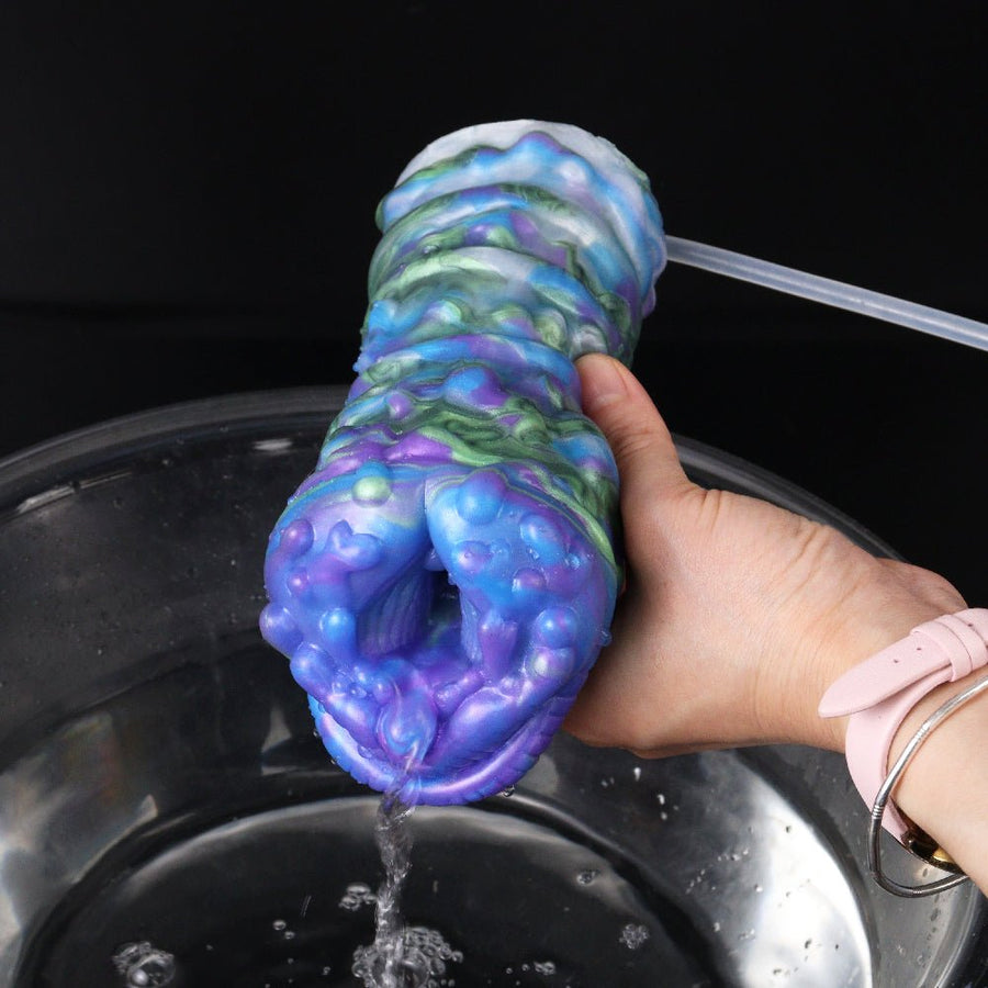 Colorful silicone stroker sleeve with long wand, held over a black container with tube attached