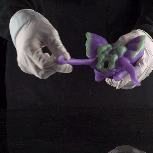 Purple alien figurine held by white gloves, facing right with curled limbs
