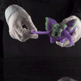 Purple alien figurine held by white gloves, facing right with curled limbs