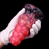 Alien butt plug held by white-gloved hand; glossy pink with purple ribbed detail