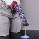 Squirting-Dildo gehalten, lila Oberfläche mit rotem Akzent, weißer Flüssigkeitsstrahl aus der Spitze